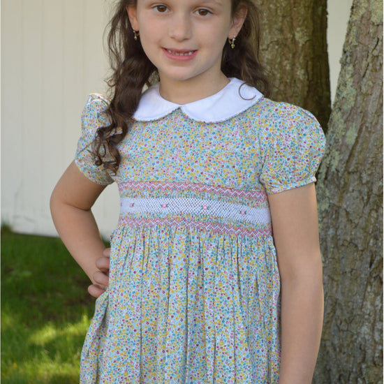 A baby girl wearing a short-sleeved green floral dress with smocked detailing and a Peter Pan collar, standing outdoors.