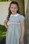 A baby girl wearing a short-sleeved green floral dress with smocked detailing and a Peter Pan collar, standing outdoors.