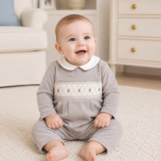 Baby in a gray outfit with white lace details sitting on a carpeted floor.