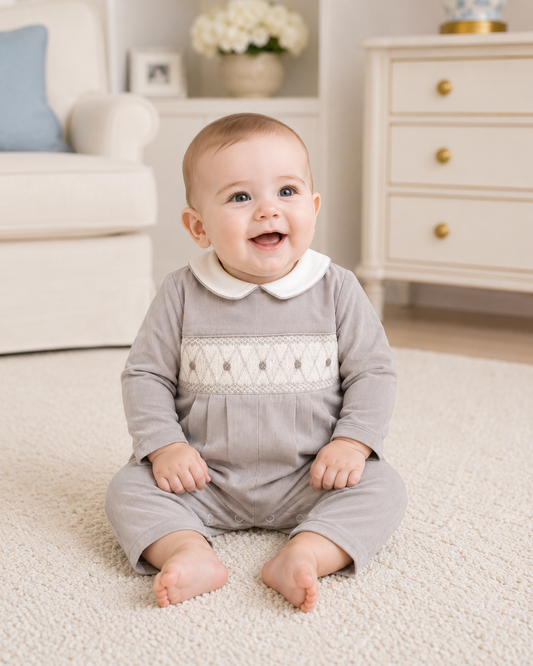 Baby in a gray outfit with white lace details sitting on a carpeted floor.