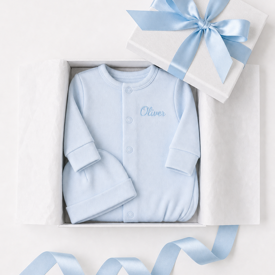 Blue baby outfit with 'Oliver' embroidered, in a white box with a blue ribbon on a light gray background