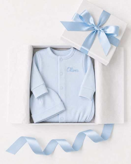 Blue baby outfit with 'Oliver' embroidered, in a white box with a blue ribbon on a light gray background