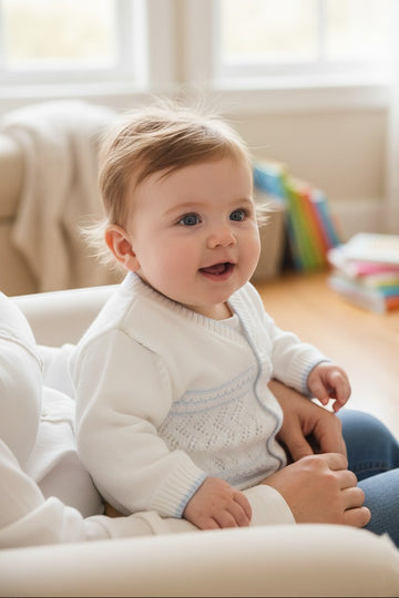 Baby sitting on a couch wearing a white sweater with colorful books in the background.
