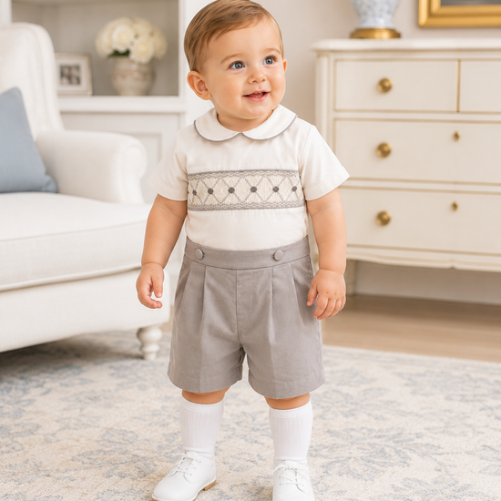Baby in a white shirt with embroidery and gray shorts standing in a living room.
