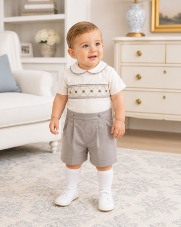 Baby in a white shirt with embroidery and gray shorts standing in a living room.