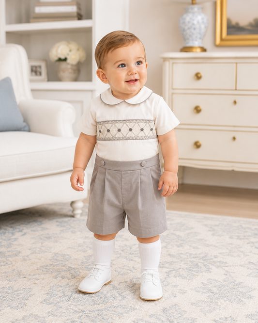 Baby in a white shirt with embroidery and gray shorts standing in a living room.