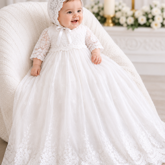 Baby in a white lace dress and bonnet sitting on a couch with a religious setting in the background.
