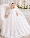 Baby in a white lace dress and bonnet sitting on a couch with a religious setting in the background.