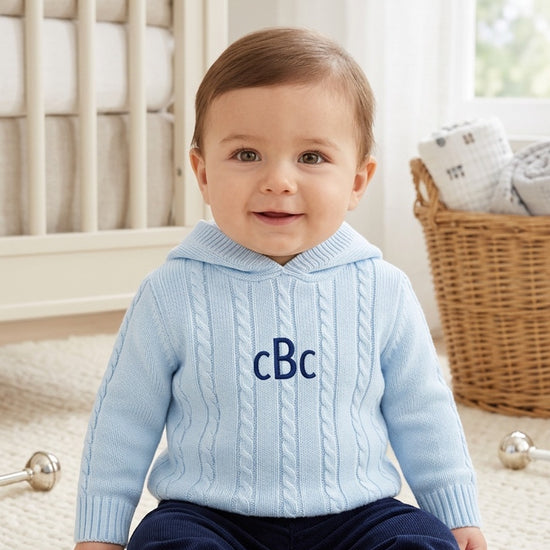 Carriage Boutique baby blue cable knit hooded sweater with navy monogram on chest worn by a smiling baby boy