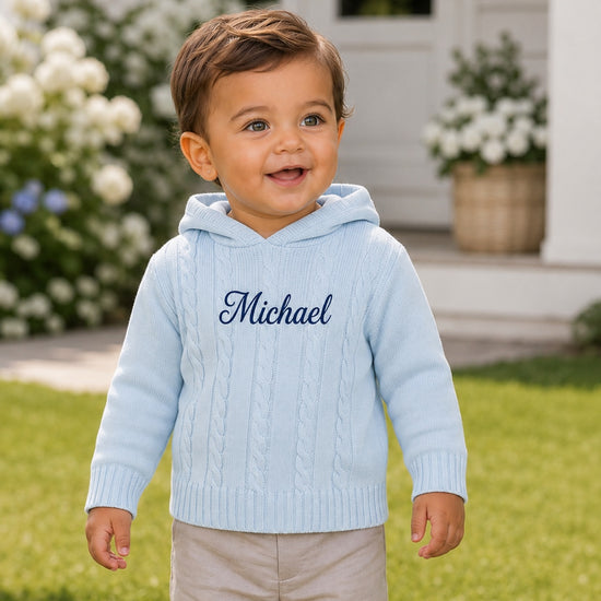 Carriage Boutique baby blue cable knit hooded sweater with custom monogram embroidery on a smiling toddler outdoors.