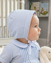 Carriage Boutique baby boy wearing a light blue shadow-patterned bonnet and matching pleated romper in a nursery.