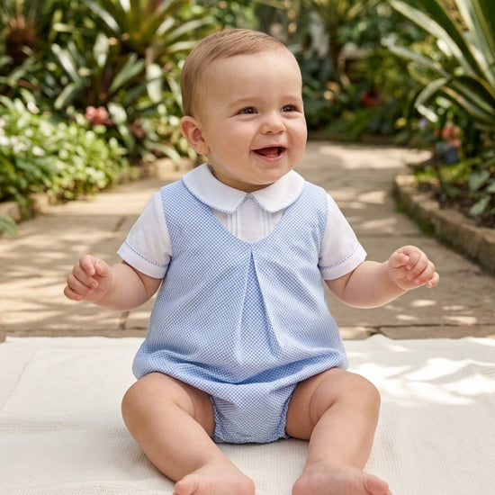 Carriage Boutique baby boy wearing a blue and white checkered bubble romper with a Peter Pan collar in a garden.