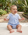 Carriage Boutique baby boy wearing a blue and white checkered bubble romper with a Peter Pan collar in a garden.