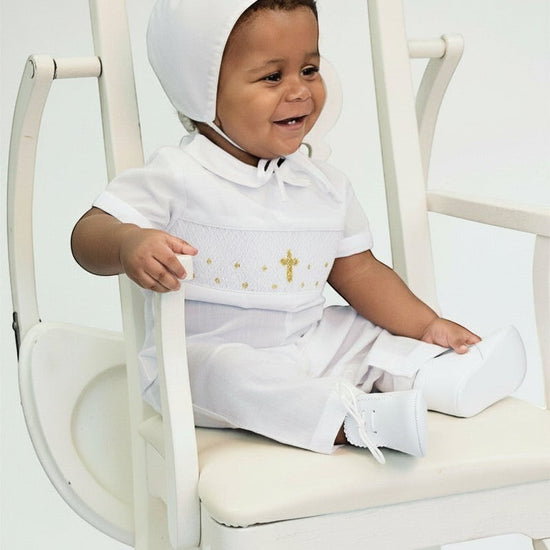 Carriage Boutique baby boy wearing a white hand-smocked longall with a gold cross and matching bonnet in a chair.