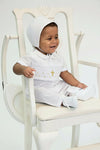 Carriage Boutique baby boy wearing a white hand-smocked longall with a gold cross and matching bonnet in a chair.
