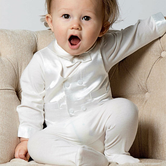 Carriage Boutique baby boy wearing a white silk vest and cotton footsie christening outfit sitting in a chair.