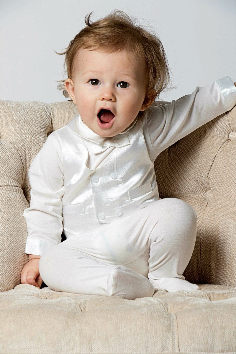 Carriage Boutique baby boy wearing a white silk vest and cotton footsie christening outfit sitting in a chair.