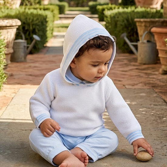 Carriage Boutique baby boy wearing a white hooded cotton sweater with blue striped trim, sitting on a stone path.