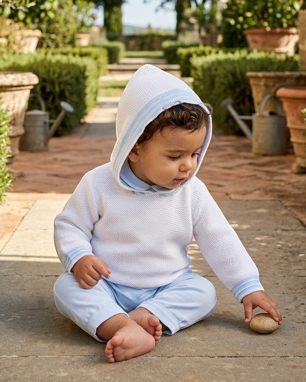 Carriage Boutique baby boy wearing a white hooded cotton sweater with blue striped trim, sitting on a stone path.