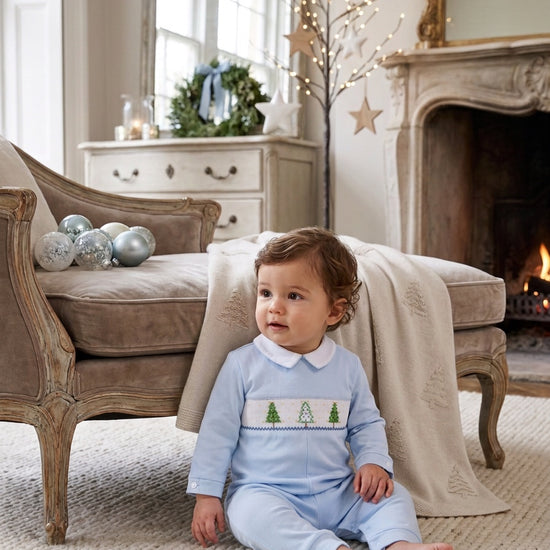 Carriage Boutique baby boy wearing a light blue longall with smocked Christmas tree embroidery in a festive room