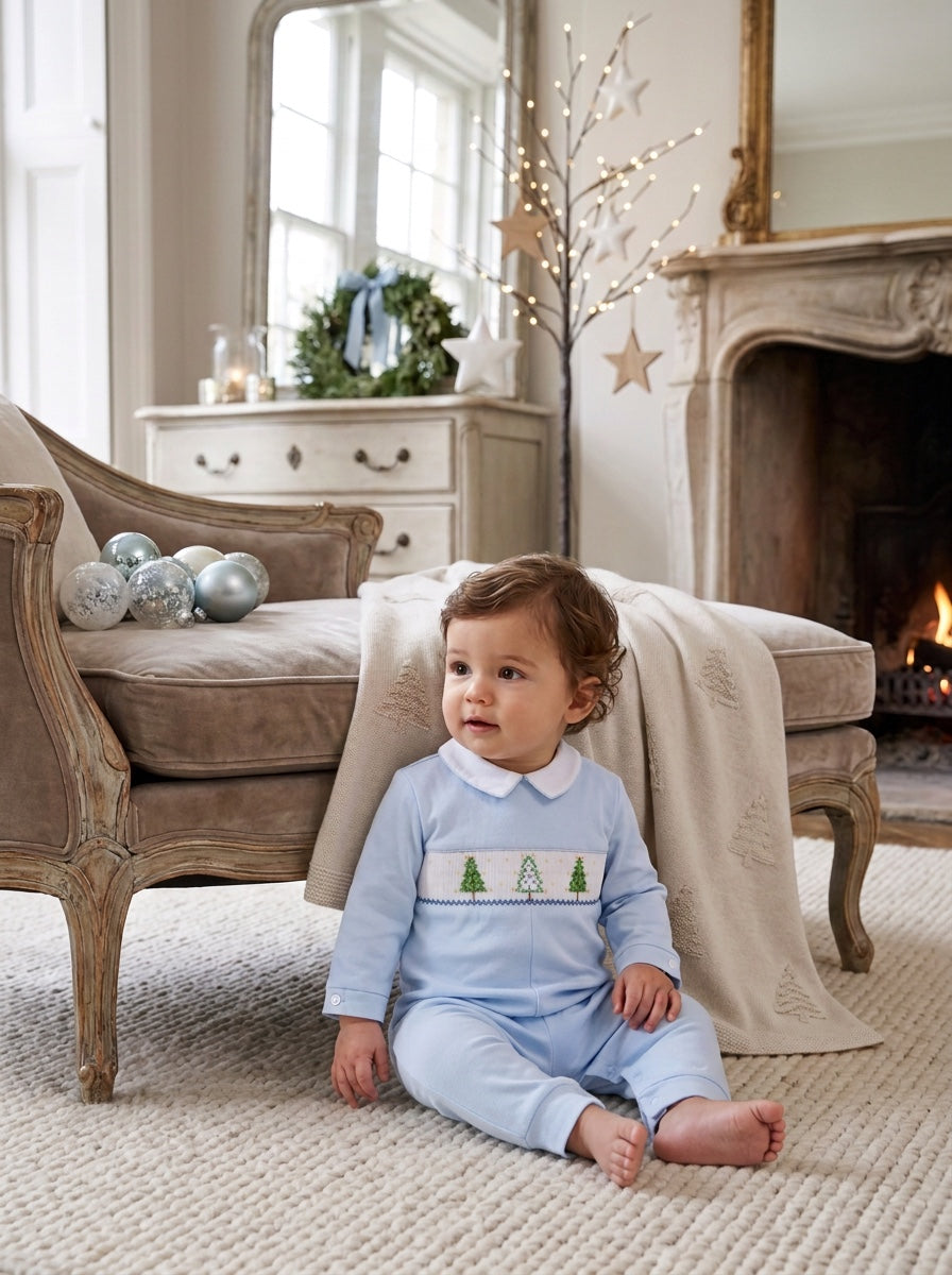 Carriage Boutique baby boy wearing a light blue longall with smocked Christmas tree embroidery in a festive room