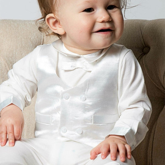 Carriage Boutique baby boy wearing a white christening footsie and silk vest sitting in a beige armchair.