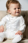 Carriage Boutique baby boy wearing a white christening footsie and silk vest sitting in a beige armchair.