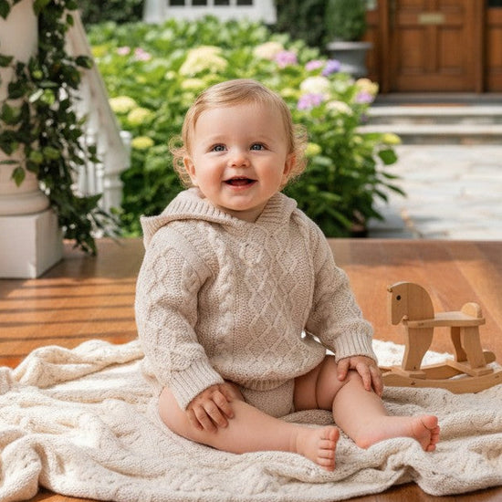 Carriage Boutique baby boy wearing a beige fisherman knit sweater with a hood, sitting on a porch with a toy horse.