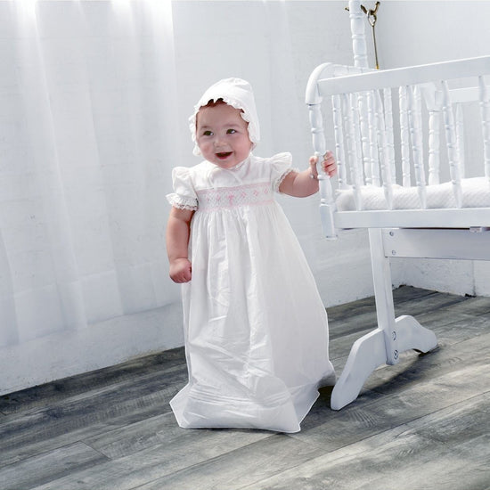 Carriage Boutique baby girl in a white christening gown with pink smocking, embroidered cross, and matching bonnet.