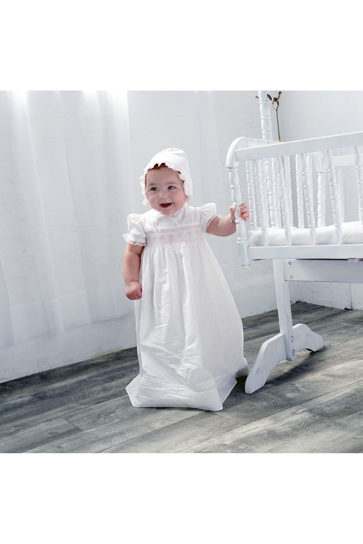 Carriage Boutique baby girl in a white christening gown with pink smocking, embroidered cross, and matching bonnet.
