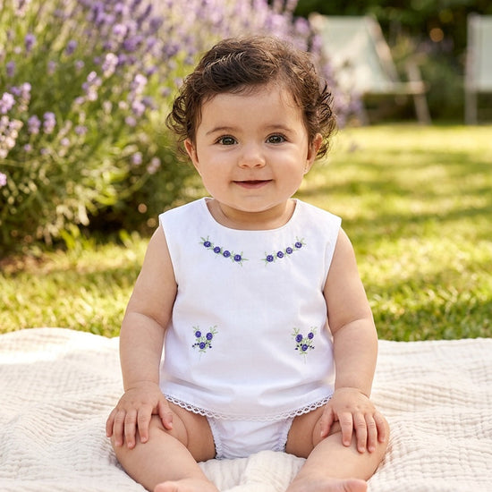Baby girl wearing a Carriage Boutique white cotton sleeveless diaper set with lilac flower bullion embroidery.