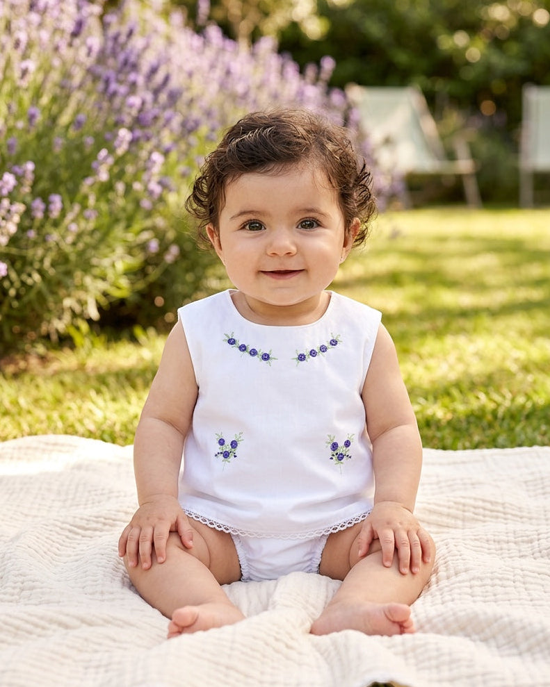 Baby girl wearing a Carriage Boutique white cotton sleeveless diaper set with lilac flower bullion embroidery.