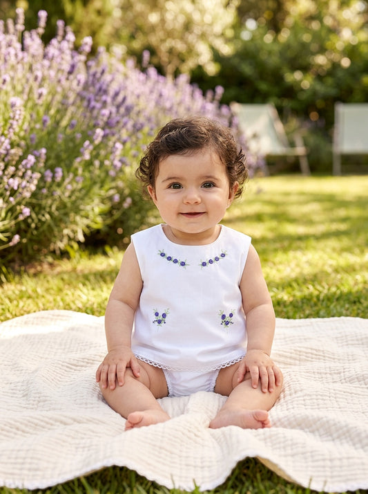 Baby girl wearing a Carriage Boutique white cotton sleeveless diaper set with lilac flower bullion embroidery.
