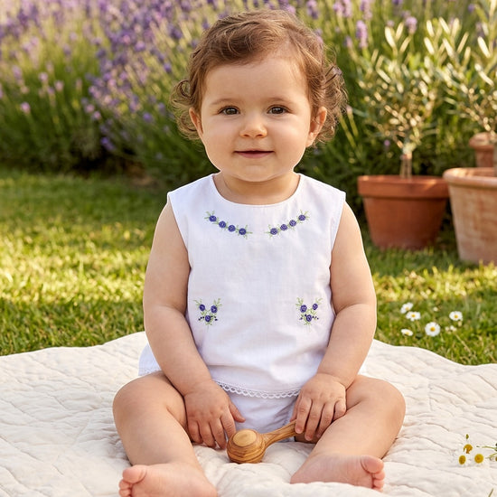 Carriage Boutique baby girl sleeveless cotton diaper set in white with lilac flower bullion embroidery in a garden.