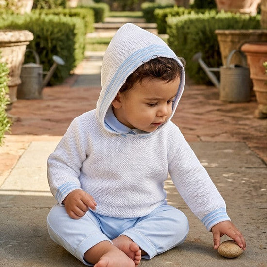 Carriage Boutique baby boy wearing a white hooded cotton sweater with blue striped trim, sitting on a stone path.