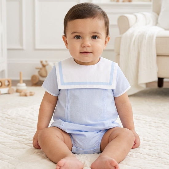 Carriage Boutique baby boy bubble romper in light blue and white with a sailor-style collar, sitting on a rug.