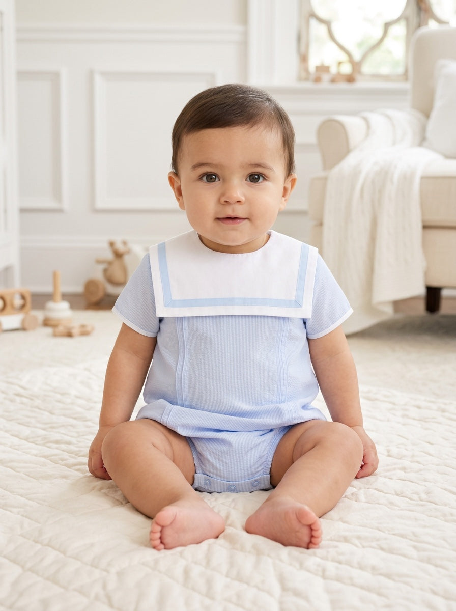 Carriage Boutique baby boy bubble romper in light blue and white with a sailor-style collar, sitting on a rug.
