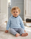 Carriage Boutique baby boy wearing a light blue hooded cotton sweater with a zip back detail in a nursery setting.