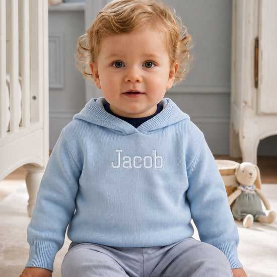 Carriage Boutique blue cotton hooded sweater with Jacob embroidery worn by a toddler sitting in a nursery
