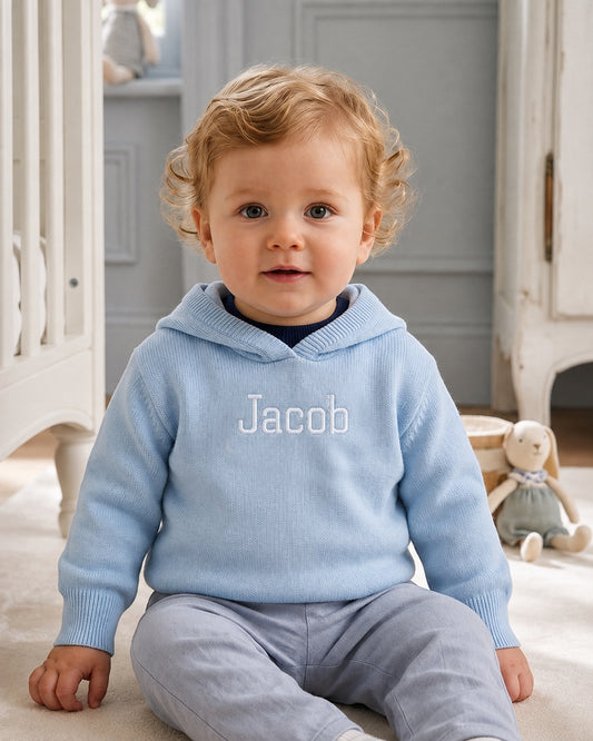 Carriage Boutique blue cotton hooded sweater with Jacob embroidery worn by a toddler sitting in a nursery