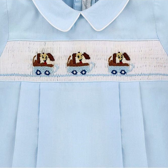Carriage Boutique light blue cotton baby boy romper with hand-smocked white panel featuring brown dogs in blue wagons.
