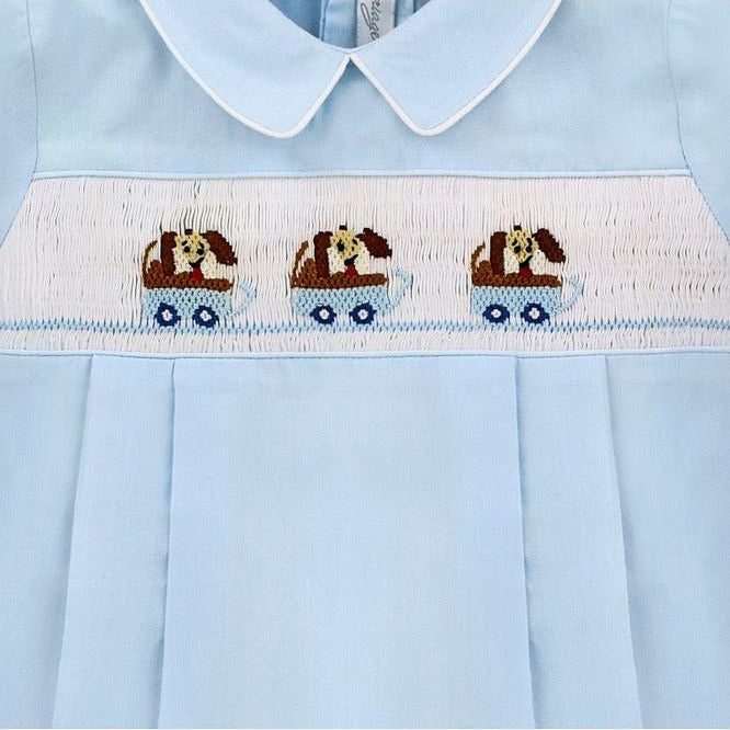 Carriage Boutique light blue cotton baby boy romper with hand-smocked white panel featuring brown dogs in blue wagons.