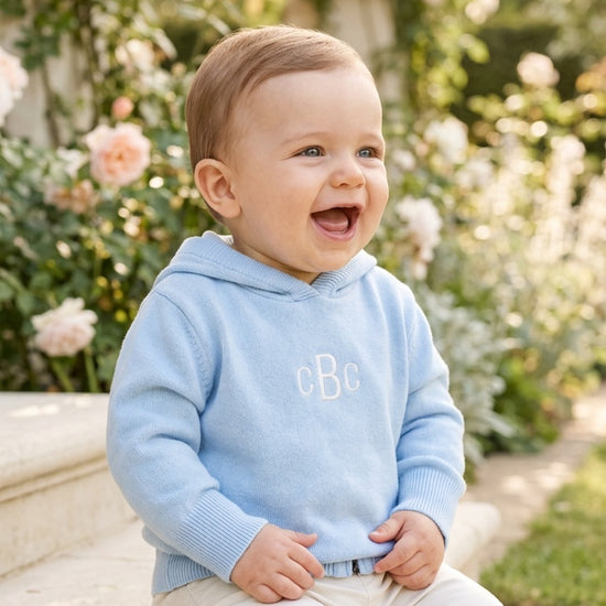 Carriage Boutique baby boy wearing a soft blue cotton hooded sweater with monogram detail in a garden setting