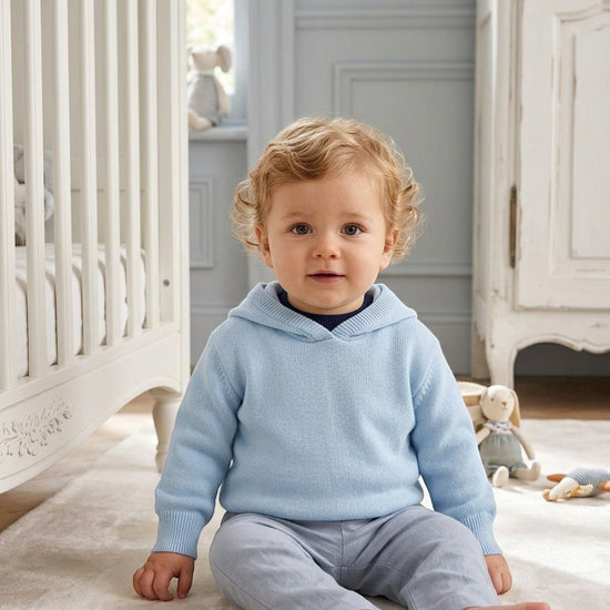 Carriage Boutique baby boy wearing a soft blue hooded cotton sweater with a zip back detail in a nursery setting.