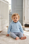 Carriage Boutique baby boy wearing a soft blue hooded cotton sweater with a zip back detail in a nursery setting.