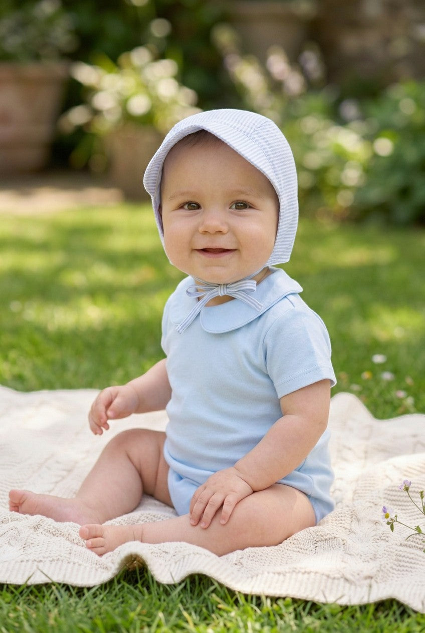 Baby boy wearing a Carriage Boutique blue seersucker sun hat and light blue cotton onesie sitting on a blanket.