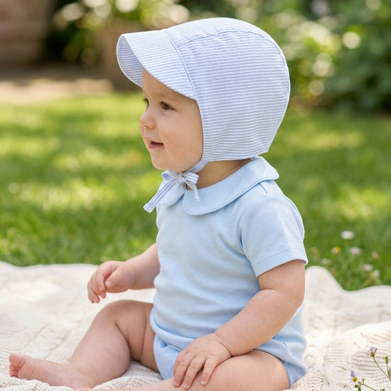 Carriage Boutique baby boy wearing a blue seersucker sun hat with chin ties while sitting on a blanket outdoors.