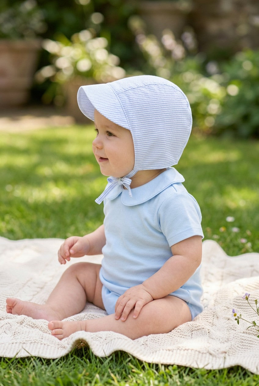 Carriage Boutique baby boy wearing a blue seersucker sun hat with chin ties while sitting on a blanket outdoors.
