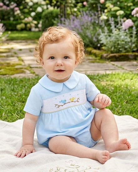 Carriage Boutique baby boy wearing a light blue hand-smocked romper with bird embroidery sitting in a garden.