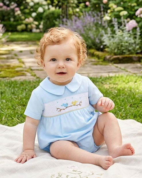 Carriage Boutique baby boy wearing a light blue hand-smocked romper with bird embroidery sitting in a garden.
